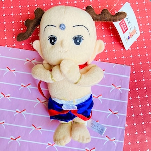 May include: A plush doll of a cartoon character with a bald head, large eyes, and small antlers. The doll wears blue shorts, a red collar, and gold armbands. The doll is on a pink and red patterned surface.