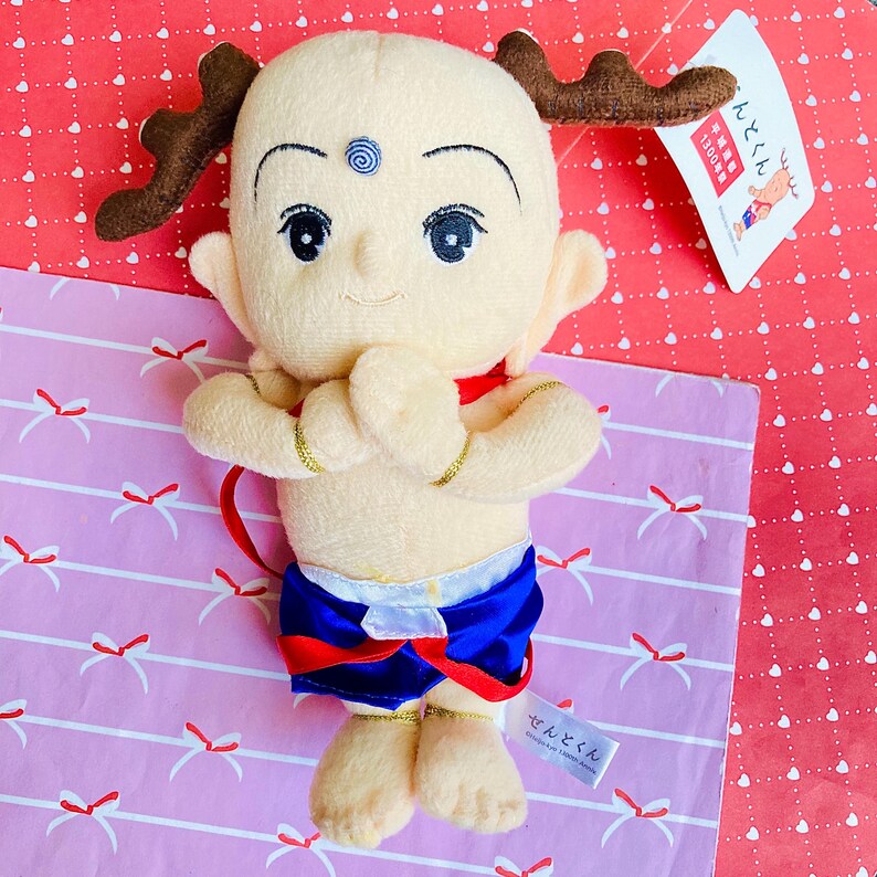 May include: A plush doll of a cartoon character with a bald head, large eyes, and small antlers. The doll wears blue shorts, a red collar, and gold armbands. The doll is on a pink and red patterned surface.