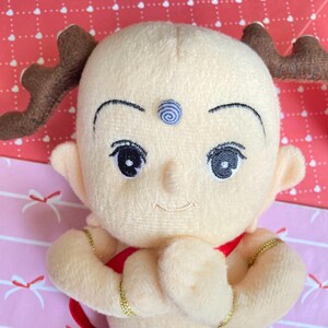 May include: A plush doll with a light yellow complexion, large black eyes, and a small smile. The doll has brown antler-like appendages and wears a red garment. Gold-colored bracelets adorn its wrists. A blue spiral design is on its forehead.