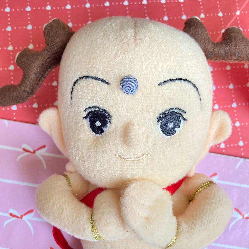 May include: A plush doll with a light yellow complexion, large black eyes, and a small smile. The doll has brown antler-like appendages and wears a red garment. Gold-colored bracelets adorn its wrists. A blue spiral design is on its forehead.