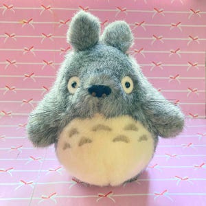 Puede incluir: Un peluche gris de Totoro con una barriga color crema y marcas distintivas. El juguete tiene ojos grandes y redondos, una pequeña nariz negra y bigotes. El fondo es una superficie rosa con un patrón repetido de lazos blancos.
