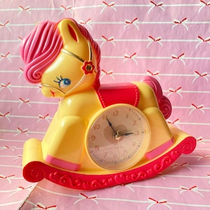 May include: A yellow rocking horse alarm clock with a pink mane and a red saddle. The clock face has black hands and white numbers.