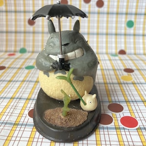 May include: A gray and cream-colored figurine of a round creature with pointed ears and a wide smile, holding a black umbrella. The figure stands on a dark oval base with a small green sprout in soil and a tiny white creature beside it.