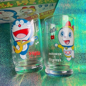 Puede incluir: Dos vasos de vidrio transparente con personajes de dibujos animados. Uno tiene un gato azul tocando la guitarra, el otro un gato amarillo sosteniendo una flor. Los vasos tienen texto japon&eacute;s y las palabras "mitsuya & Bireley's".