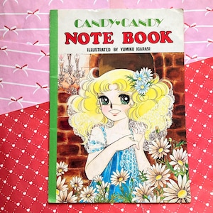 Può includere: Un quaderno vintage "Candy Candy" con dorso verde. La copertina presenta un'illustrazione manga di una ragazza bionda con fiori blu tra i capelli, circondata da margherite. Il testo recita "Candy Candy Note Book".