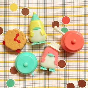 Puede incluir: Una colección de figuras coloridas y caprichosas, que incluyen un pingüino amarillo con un sombrero verde, un pingüino rosa con un sombrero blanco, un reloj con forma de galleta y un juguete de color turquesa y coral. El fondo es un patrón de cuadros con lunares.