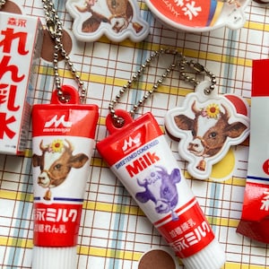 May include: A collection of red and white tube-shaped keychains and cow-shaped charms. The keychains feature the text "Morinaga" and Japanese text, with a cow illustration. The charms also feature cow illustrations. The items are on a patterned surface.
