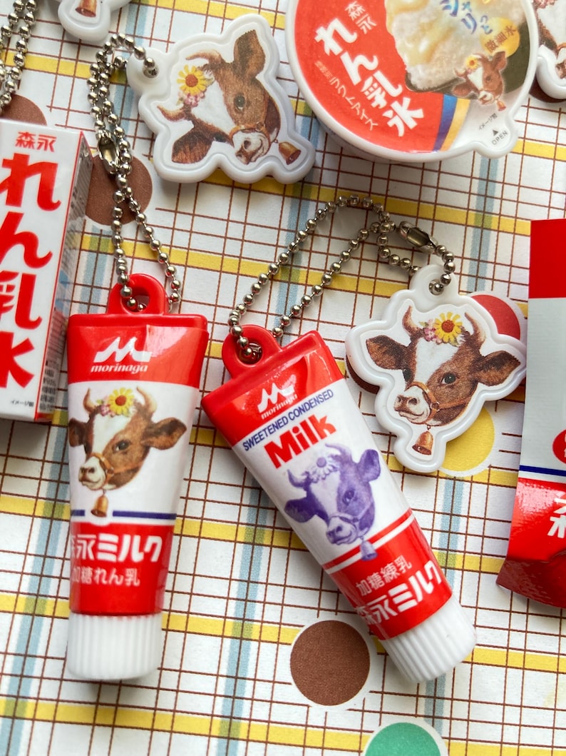 May include: A collection of red and white tube-shaped keychains and cow-shaped charms. The keychains feature the text "Morinaga" and Japanese text, with a cow illustration. The charms also feature cow illustrations. The items are on a patterned surface.