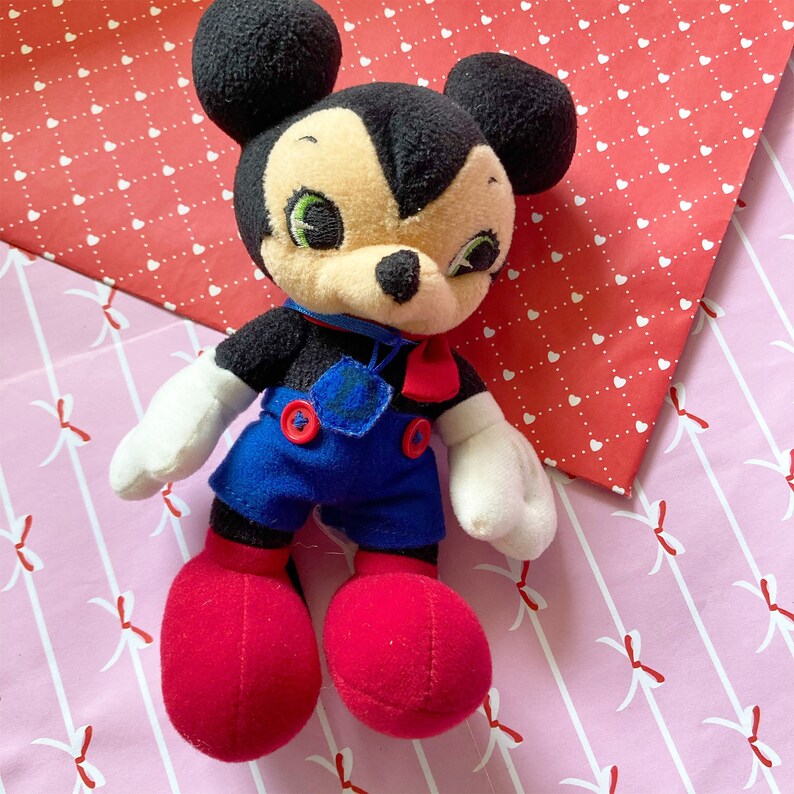 May include: A vintage plush toy of a cartoon mouse character. The toy has black fur, a cream-colored face, and large black ears. It wears blue overalls with red buttons, red shoes, and white gloves. A red bow tie and blue collar complete the outfit.