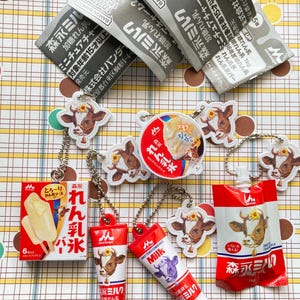 May include: A collection of miniature Japanese food items, including ice cream bars, milk cartons, and milk tube keychains. The items feature cow illustrations and Japanese text. Some items are packaged in red and white containers.