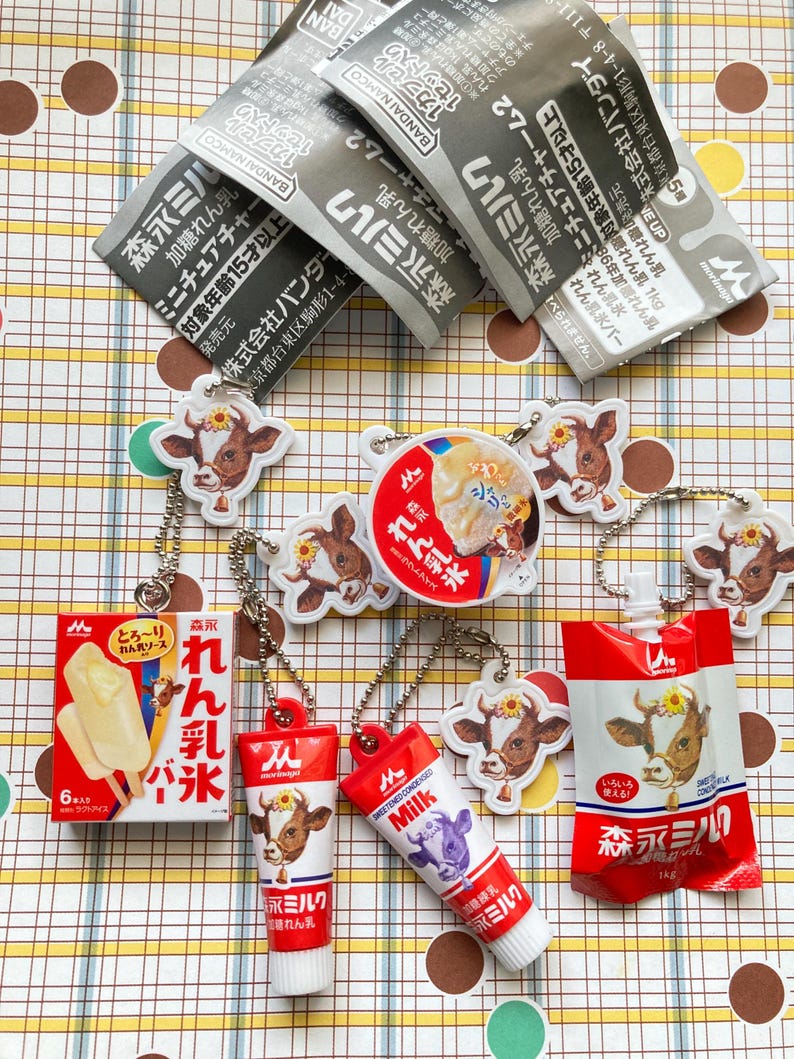 May include: A collection of miniature Japanese food items, including ice cream bars, milk cartons, and milk tube keychains. The items feature cow illustrations and Japanese text. Some items are packaged in red and white containers.