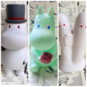 Pode incluir: Três figuras de plástico branco. A primeira é um personagem Moomin com um chapéu-coco preto, a segunda é um personagem Moomin verde segurando uma maçã vermelha, e a terceira é um par de personagens fantasmas brancos.