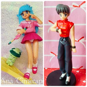 Puede incluir: Dos figuras de estilo anime. La primera es una chica con pelo azul, un atuendo rosa y una pequeña criatura verde. La segunda es una figura masculina con una parte superior roja y pantalones negros, de pie sobre una base negra.