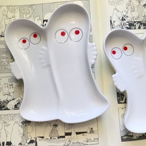 May include: Two white ceramic divided plates shaped like cartoon characters with red eyes. The plates are on a page of a book with black and white illustrations.