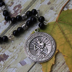 May include: A silver pendant necklace with a black beaded chain. The pendant features a circular design with the words "St. Christopher Be My Guide" and an image of St. Christopher carrying a child.