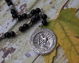 May include: A silver pendant necklace with a black beaded chain. The pendant features a circular design with the words "St. Christopher Be My Guide" and an image of St. Christopher carrying a child.