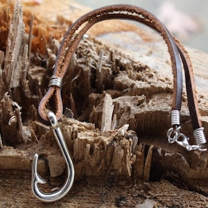 May include: A brown leather necklace with a silver-colored fish hook pendant. The leather strap is wrapped with silver wire at the ends. The hook is a shiny, curved shape.