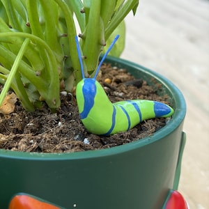 May include: A whimsical, hand-painted ceramic slug figurine in a green and blue color scheme. The slug is resting on the soil of a potted plant. The pot is dark green with a mushroom design on the side.