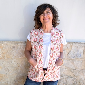 Puede incluir: Una mujer lleva una camiseta blanca y un chaleco sin mangas con un estampado floral en tonos rosa, marrón y blanco. El chaleco tiene dos bolsillos y un cierre de botón.