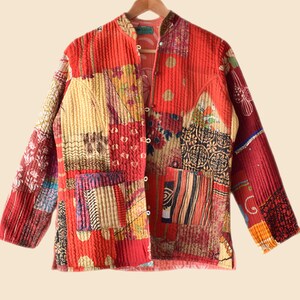 Puede incluir: Una chaqueta acolchada de retazos roja, amarilla y marrón con cierre de botón. La chaqueta tiene un cuello mandarín y mangas largas.