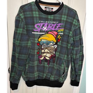 Könnte beinhalten: Ein langärmeliges Sweatshirt mit Rundhalsausschnitt in einem grün-dunkelblauen Karomuster. Die Vorderseite zeigt eine Grafik einer Cartoon-Figur, die an einen Stuhl gefesselt ist, mit dem Text "COLLECTIVELY STABLE" und "BKYS" auf dem Innenetikett.