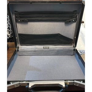 May include: An open black hardshell briefcase with a grey fabric interior lining. The interior features a black accordion-style document organizer, a black pocket, and a small Samsonite brand label. The case has a metallic silver handle.