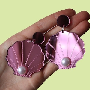 May include: Pink mirrored seashell earrings with a pearl accent. The earrings are dangling and have a shiny finish.