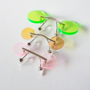 May include: Three pairs of round, colourful sunglasses with silver metal frames. The sunglasses are stacked on top of each other. The top pair is green, the middle pair is yellow, and the bottom pair is pink.