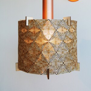 May include: A copper and wood pendant light with a geometric pattern on the shade. The light is hanging from a black cord and is attached to a copper wall mount.