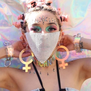 May include: A woman wearing a silver rhinestone face mask with a silver rhinestone choker necklace that says "techno". She is also wearing large gold earrings in the shape of the female symbol and has pink hair.
