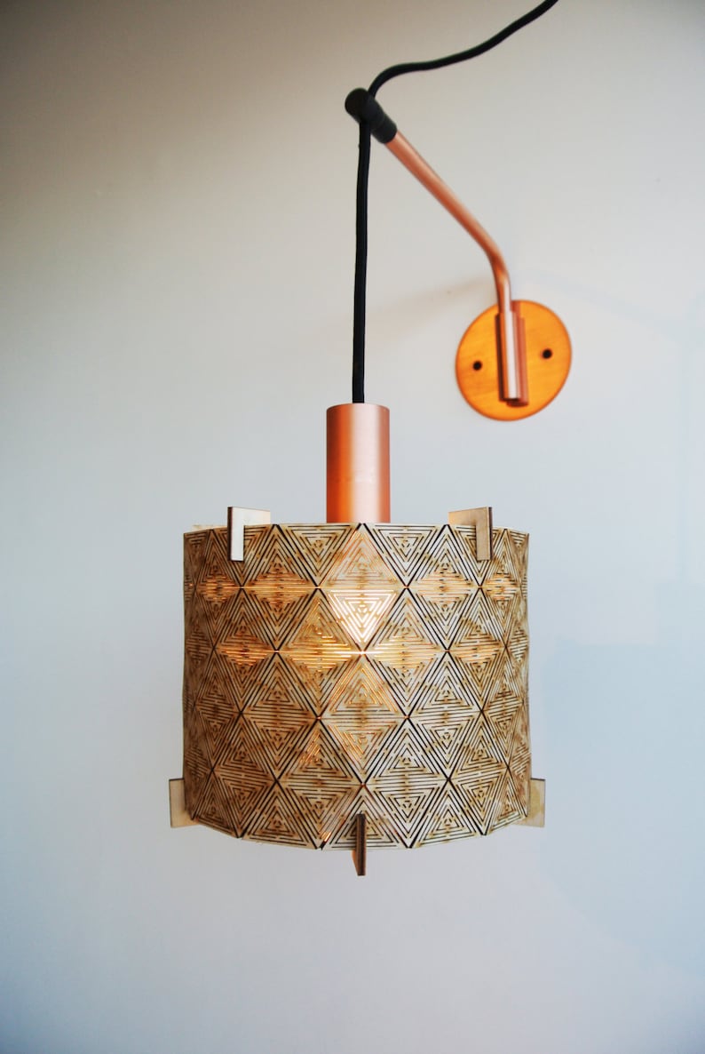 May include: A copper and wood pendant light with a geometric pattern on the shade. The light is hanging from a black cord and is attached to a copper wall mount.