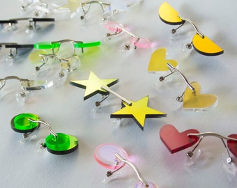Puede incluir: Una colección de gafas de colores y espejadas con diferentes formas, incluyendo estrellas, corazones y círculos. Las gafas están diseñadas para sujetarse a las gafas existentes.