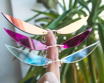 Puede incluir: Tres pares de gafas de sol con lentes de diferentes colores. Las lentes son de color azul claro, rosa y cobre. Las gafas de sol tienen todas la forma de un ala.