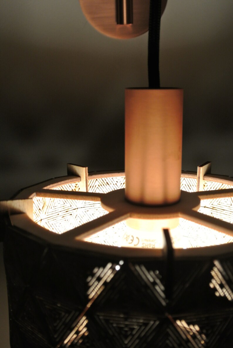 May include: A wooden pendant light with a geometric pattern and a copper finish. The light is hanging from a black cord and has a copper ceiling mount. The light is on and illuminating the pattern.