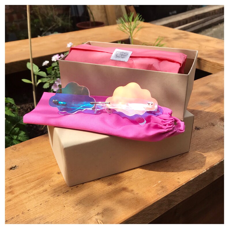 Puede incluir: Gafas de sol en forma de nube con lentes iridiscentes azules y rosas. Las gafas est&aacute;n en una bolsa rosa y una caja beige.