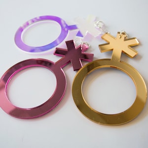 May include: Three mirrored acrylic gender symbols. One is gold, one is pink, and one is purple.