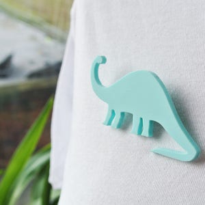 Pode incluir: Um broche em forma de dinossauro azul claro em uma camiseta branca.