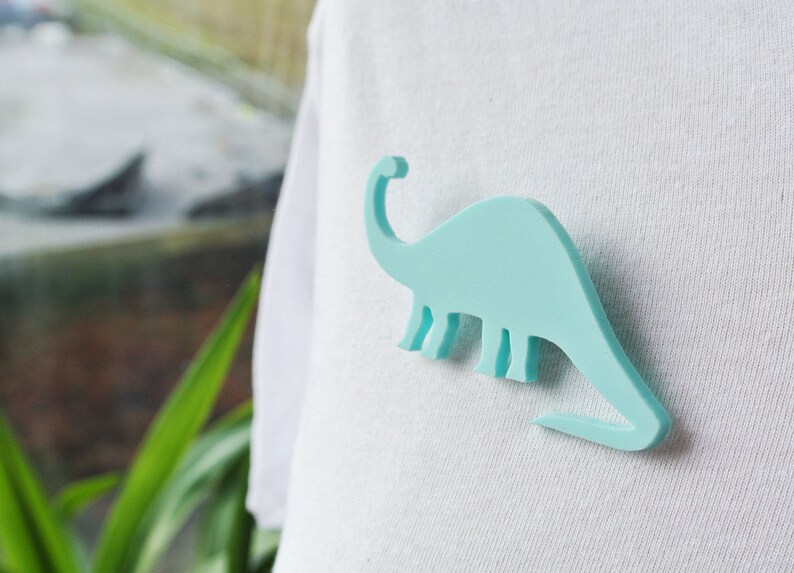 Pode incluir: Um broche em forma de dinossauro azul claro em uma camiseta branca.