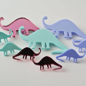 Pode incluir: Um conjunto de sete brincos de acr&iacute;lico em forma de dinossauro em v&aacute;rias cores, incluindo rosa, azul e turquesa. Os brincos s&atilde;o de diferentes tamanhos e formas, e alguns t&ecirc;m um acabamento espelhado.