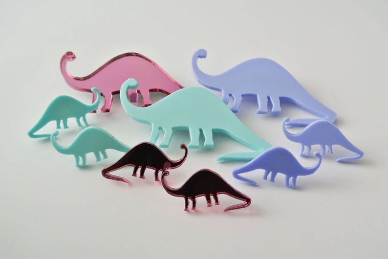 Pode incluir: Um conjunto de sete brincos de acr&iacute;lico em forma de dinossauro em v&aacute;rias cores, incluindo rosa, azul e turquesa. Os brincos s&atilde;o de diferentes tamanhos e formas, e alguns t&ecirc;m um acabamento espelhado.