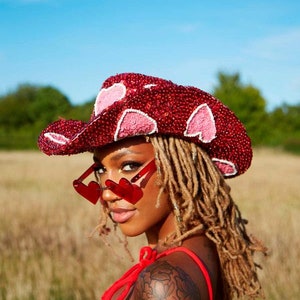 May include: A woman wearing a red sequined cowboy hat with pink hearts, red heart-shaped sunglasses, and a red sequined top with pink hearts. She has long blonde dreadlocks and a black floral tattoo on her arm.