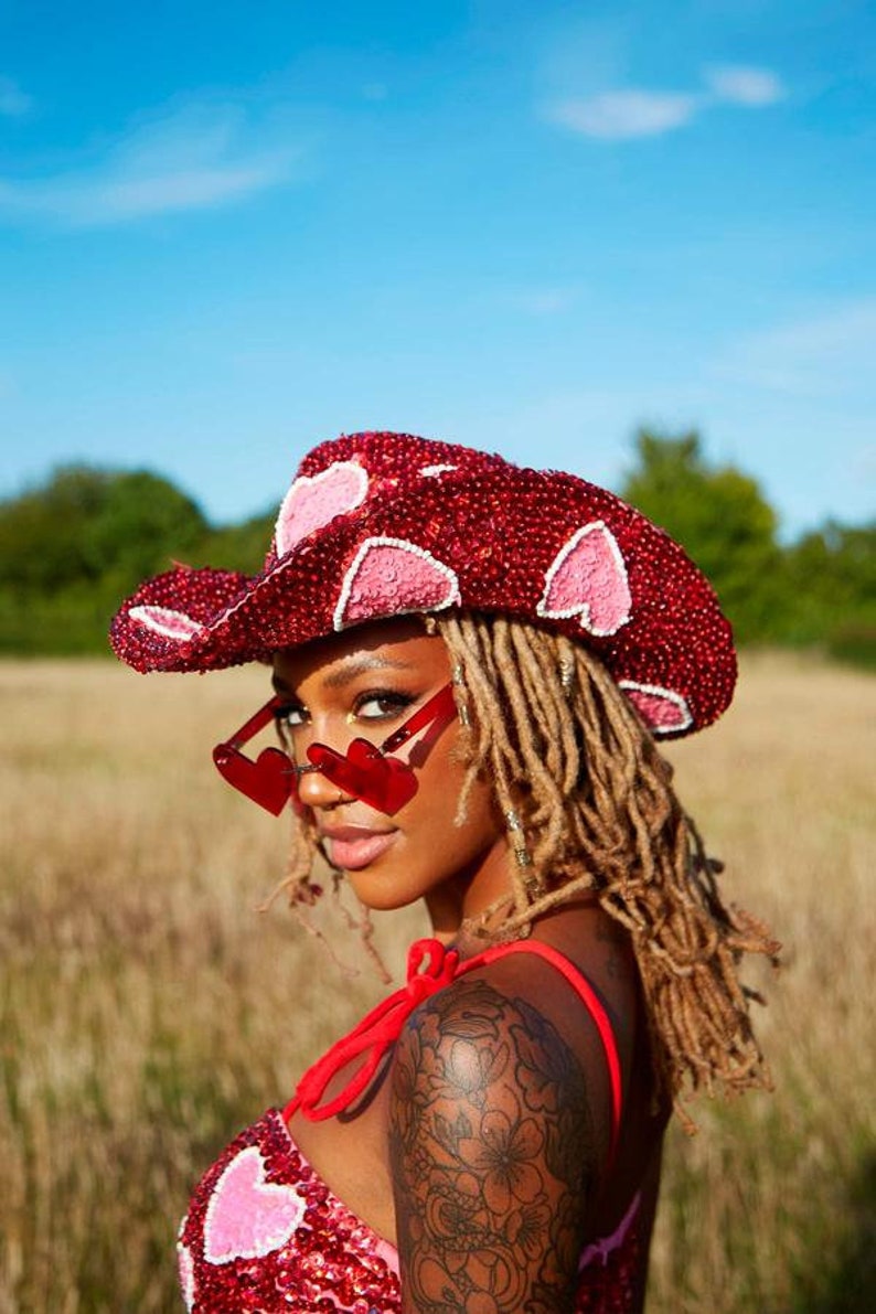 May include: A woman wearing a red sequined cowboy hat with pink hearts, red heart-shaped sunglasses, and a red sequined top with pink hearts. She has long blonde dreadlocks and a black floral tattoo on her arm.