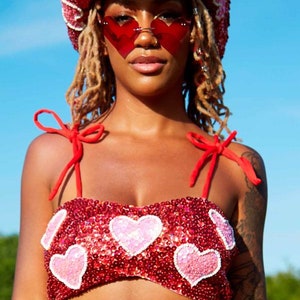 May include: A red sequined crop top with pink heart embellishments and red straps. The top is worn with a red sequined cowboy hat with pink hearts and red heart-shaped sunglasses.