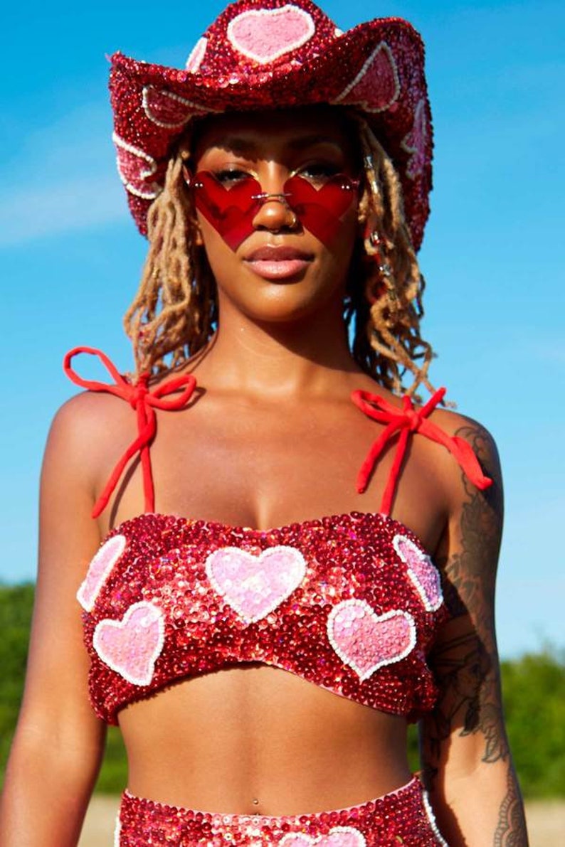May include: A red sequined crop top with pink heart embellishments and red straps. The top is worn with a red sequined cowboy hat with pink hearts and red heart-shaped sunglasses.