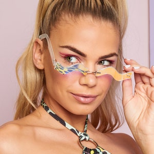 May include: A woman wearing a green and black leopard print halter top with a silver ring accent. She is wearing clear, iridescent sunglasses with a wavy, abstract design.