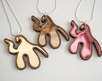 Puede incluir: Tres collares con colgantes de madera abstractos. Los colgantes están hechos de madera con un acabado espejado. Los colgantes son de color dorado, oro rosa y rosa.