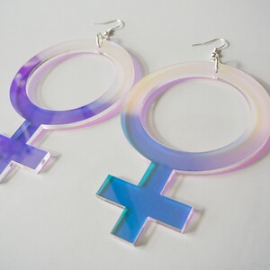 May include: A pair of iridescent acrylic earrings shaped like the female gender symbol. The earrings are large and have a holographic finish.