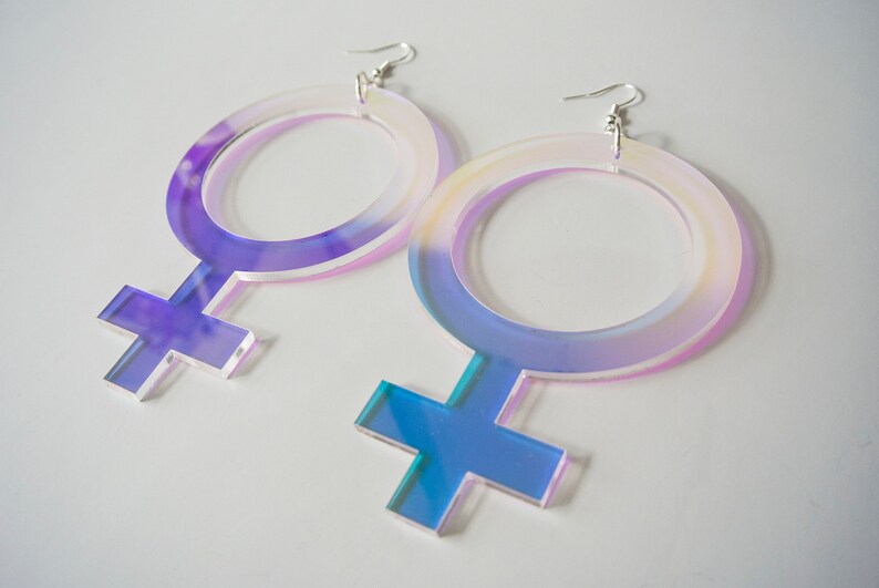 May include: A pair of iridescent acrylic earrings shaped like the female gender symbol. The earrings are large and have a holographic finish.
