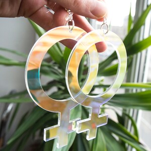 May include: A pair of iridescent clear acrylic earrings shaped like the female gender symbol.