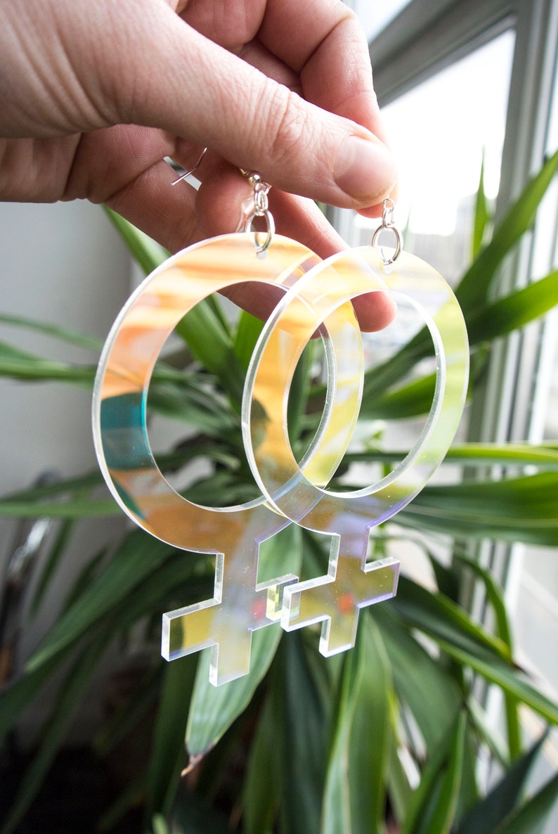 May include: A pair of iridescent clear acrylic earrings shaped like the female gender symbol.